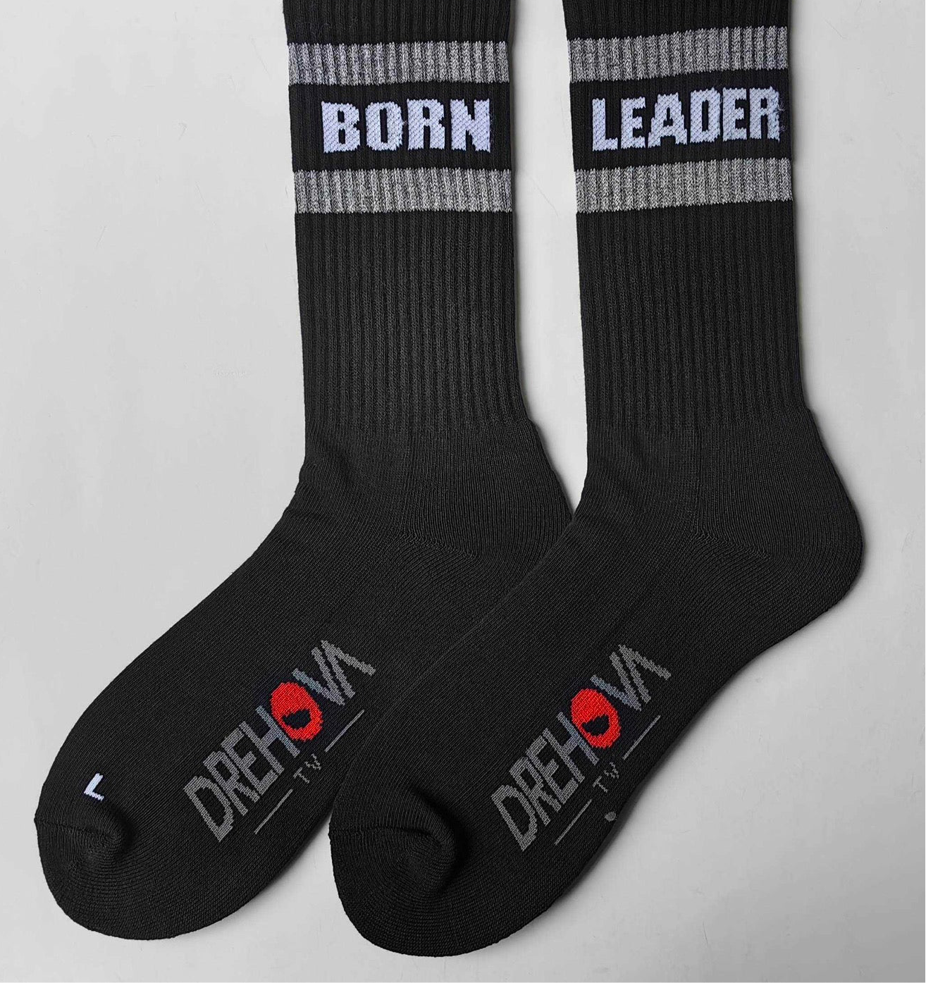 Unisex black crew socks featuring a bold, reflective design. The left sock is labeled ‘BORN’ and the right sock is labeled ‘LEADER.’ The socks also showcase the DREHOVA TV logo on the foot in a sleek combination of grey and red. These socks are perfect for individuals who embrace leadership and individuality. Available in men’s shoe sizes 6-12.